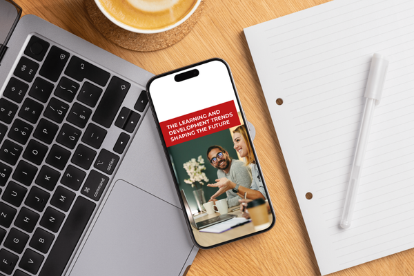 Mock-up of iPhone on a desk with the 2025 Learning and Development Trends report cover, alongside a laptop, coffee cup, and notebook